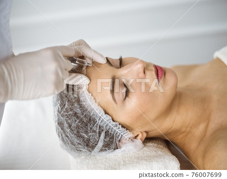 Beautician doing beauty procedure with syringe to face of young brunette woman in sunny clinic. Cosmetic medicine and surgery, beauty injections 76007699
