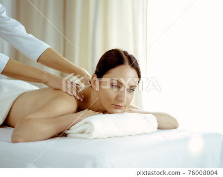 Pretty brunette woman enjoying procedure of back massage in spa salon. Beauty concept 76008004