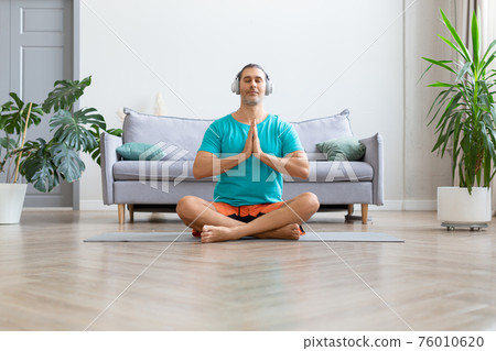Workout at home. Middle aged man in white headphones meditating while doing yoga. Prayer pose. Workout at home. Middle aged man in white headphones meditating while doing yoga. Prayer pose. 76010620