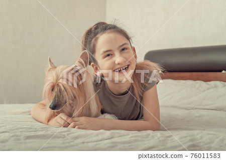 Cute smiling caucasian girl is laying on a bed hugging her cute pet. Chinese crested, hairless dog, family friend, indoor activity, portrait. Front view. 76011583