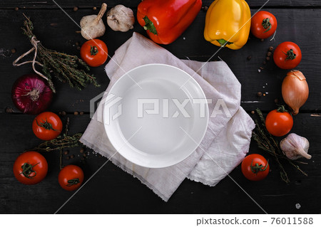 White clean plate on a napkin, bell peppers, tomatoes, garlic and onions are laying on a dark wooden table. Contrast bright colored flatlay. Top view. White clean plate on a napkin, bell peppers, tomatoes, garlic and onions are laying on a dark wooden table. Contrast bright colored flatlay. Top view. 76011588