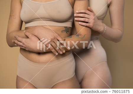 Freckled girl and tattooed girl embracing while posing on beige background 76011875