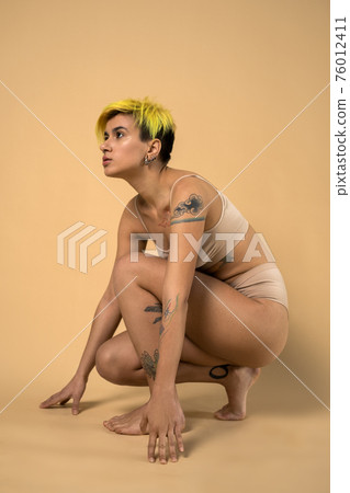 Tattooed woman with stretch marks at the body sitting while posing 76012411