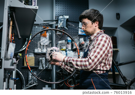 Technical expertise taking care bicycle shop. Handsome young mechanic fixing cycle wheel in workshop. Handsome repairman in workwear serving mountain bicycle. Male engineer adjusting velocipede 76013916