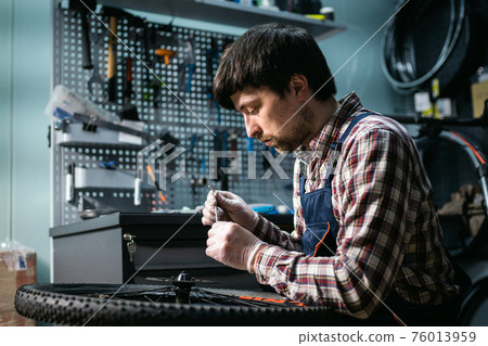 Technical expertise taking care bicycle shop. Handsome young mechanic fixing cycle wheel in workshop. Handsome repairman in workwear serving mountain bicycle. Male engineer adjusting velocipede Technical expertise taking care bicycle shop. Handsome young mechanic fixing cycle wheel in workshop. Handsome repairman in workwear serving mountain bicycle. Male engineer adjusting velocipede 76013959