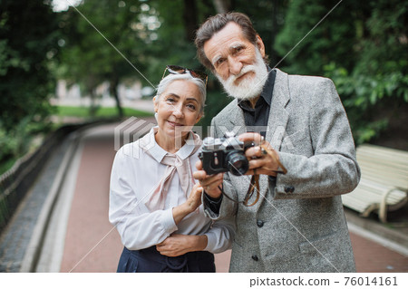 Elder couple posing at park with retro camera in hands Elder couple posing at park with retro camera in hands 76014161