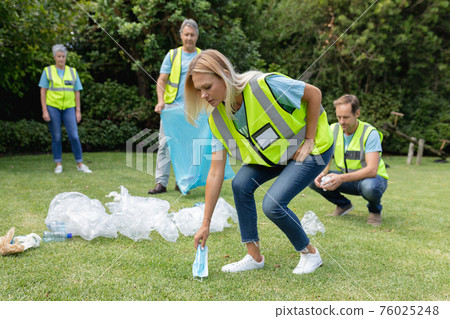 Caucasian woman picking up face mask with group of men and women collecting rubbish in field behind 76025248