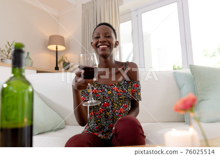 African american woman sitting on couch making video call holding glass of wine and smiling 76025514