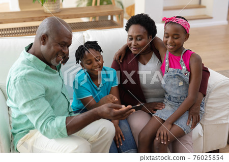 Happy african american grandfather and grandmother on couch with grandchildren looking at smartphone Happy african american grandfather and grandmother on couch with grandchildren looking at smartphone 76025854