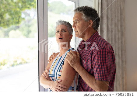 Concerned senior caucasian couple standing looking out of window embracing Concerned senior caucasian couple standing looking out of window embracing 76025881