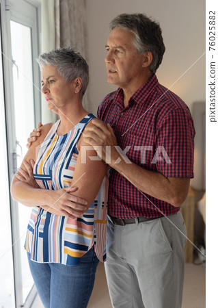 Concerned senior caucasian couple standing looking out of window embracing Concerned senior caucasian couple standing looking out of window embracing 76025882