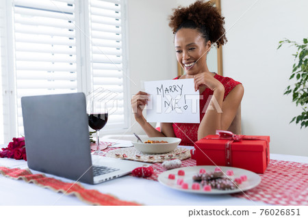 African american woman holding placard with marry me text on videocall on laptop at home 76026851