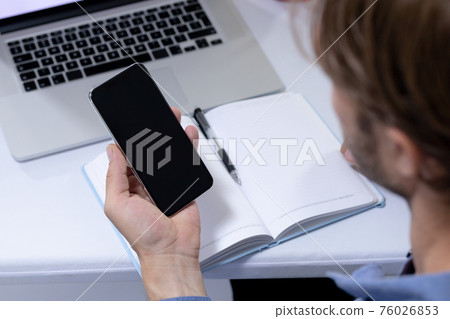 Over shoulder view of caucasian businessman sitting at desk making video call using smartphone 76026853
