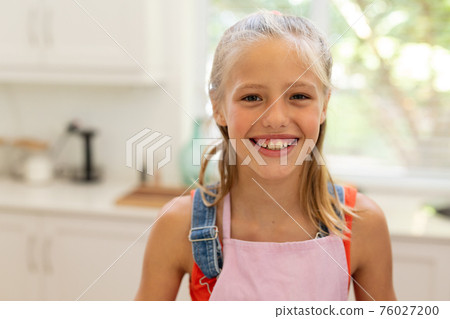 Portrait of smiling caucasian girl wearing apron standing in kitchen 76027200