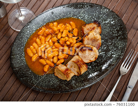 close up of fried pork with beans served on black plate on wooden table 76031100