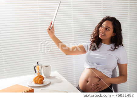 Happy pregnant young beautiful woman talking to mom using video call during morning breakfast. Concept of communication and positive attitude during pregnancy Happy pregnant young beautiful woman talking to mom using video call during morning breakfast. Concept of communication and positive attitude during pregnancy 76031497