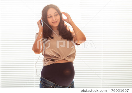 Beautiful young peaceful pregnant woman listens to pleasant classical music using smartphone and headphones. Concept of positive attitude before childbirth. Copy space Beautiful young peaceful pregnant woman listens to pleasant classical music using smartphone and headphones. Concept of positive attitude before childbirth. Copy space 76031607