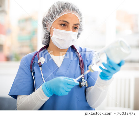 Female nurse wearing a protective mask fills a syringe with saline solution in the treatment room 76032472