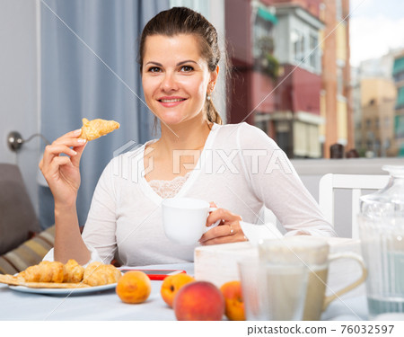 Attractive woman with tea eating croissant at table while breakfast Attractive woman with tea eating croissant at table while breakfast 76032597