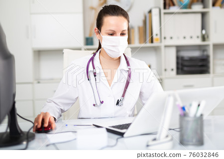Woman doctor in protective mask sitting at workplace with computer in office Woman doctor in protective mask sitting at workplace with computer in office 76032846