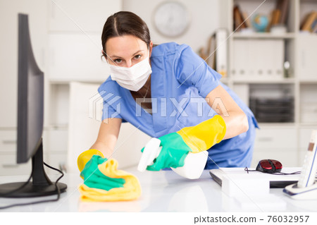 Woman in protective mask wipes dust from table in office Woman in protective mask wipes dust from table in office 76032957