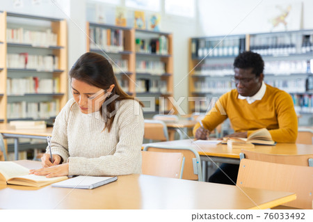 Young Latina reading books and making notes in library Young Latina reading books and making notes in library 76033492