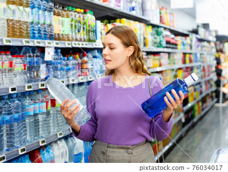 Young positive woman purchasing water in bottles in grocery store Young positive woman purchasing water in bottles in grocery store 76034017
