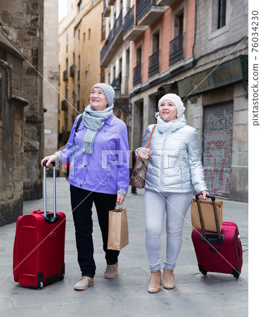 Elderly women strolling with luggage along city street 76034230