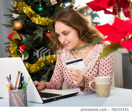 Happy young woman with credit card and laptop sitting at home against background of Christmas tree Happy young woman with credit card and laptop sitting at home against background of Christmas tree 76034381