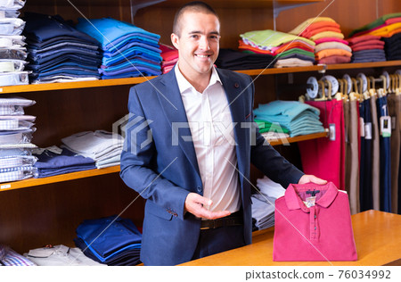 Male seller lays out shirts on shelves in clothing store 76034992