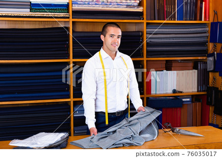 Smiling successful male administrator of clothing store standing behind counter on background of shelves with colorful garments Smiling successful male administrator of clothing store standing behind counter on background of shelves with colorful garments 76035017