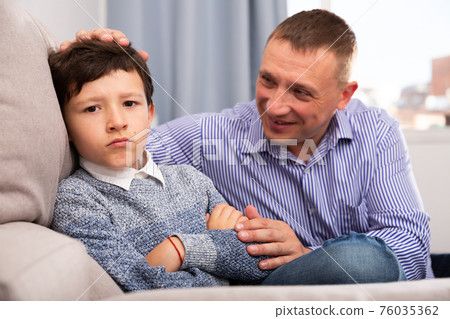 Young father consoling preteen son sitting at sofa after quarrel Young father consoling preteen son sitting at sofa after quarrel 76035362
