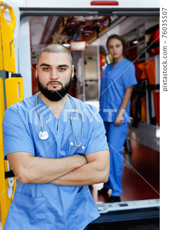 Two confident paramedicals in uniform posing near ambulance car Two confident paramedicals in uniform posing near ambulance car 76035507
