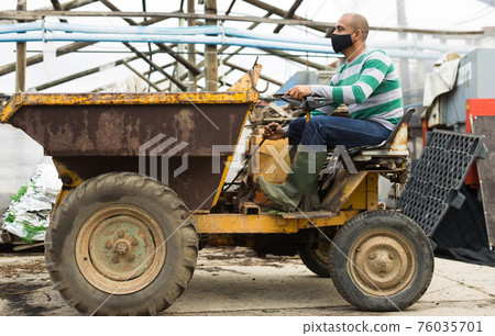 Latin american farmer wearing a protective mask during a pandemic, sits behind the wheel. 76035701