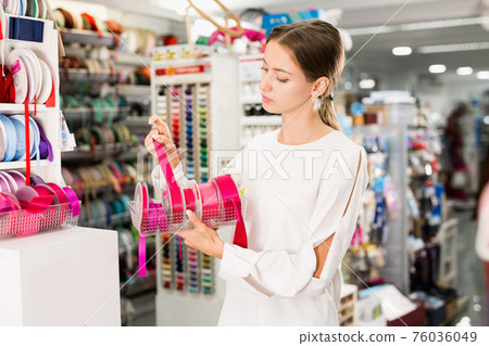 Interested young woman choosing colorful ribbons and braid for dressmaking in sewing supplies shop Interested young woman choosing colorful ribbons and braid for dressmaking in sewing supplies shop 76036049