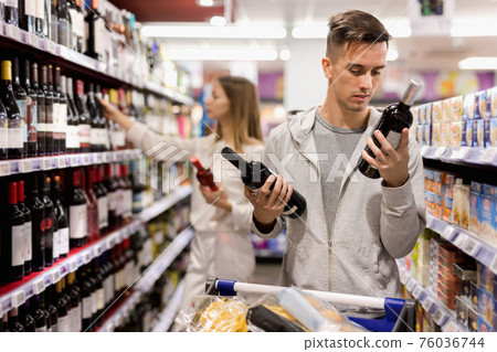 Portrait of young positive male customer selecting wine in supermarket Portrait of young positive male customer selecting wine in supermarket 76036744