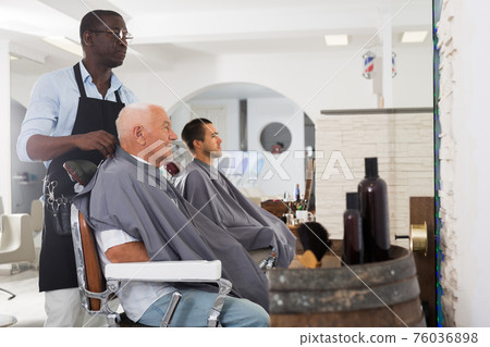 African barber putting on hairdresser cape to elderly client 76036898