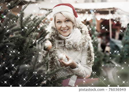 Woman in Christmas hat choosing decorations and Christmas tree Woman in Christmas hat choosing decorations and Christmas tree 76036949