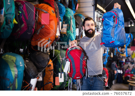 Smiling male customer examining rucksacks in store Smiling male customer examining rucksacks in store 76037672