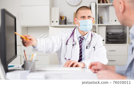 Male doctor in a protective mask consults a patient in the office Male doctor in a protective mask consults a patient in the office 76038011