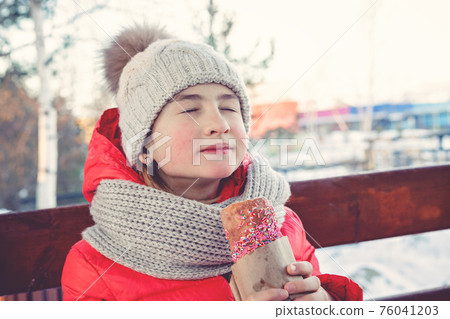 Young beautiful caucasian girl with closed eyes dressed in a bright jacket eating a colorful pie. Winter outdoor activity, happy childhood. Front view. Young beautiful caucasian girl with closed eyes dressed in a bright jacket eating a colorful pie. Winter outdoor activity, happy childhood. Front view. 76041203