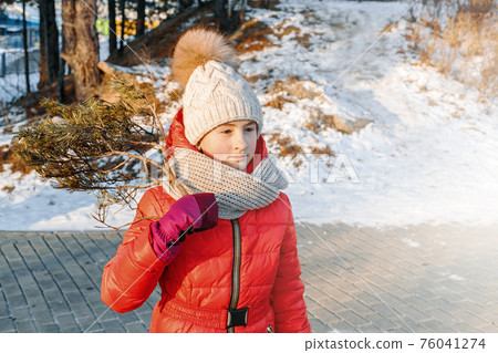 Young cute caucasian girl in bright clothes is holding a tree branch. Winter outdoor activity, happy childhood. Front view. 76041274