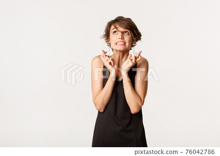 Image of hopeful woman in black dress looking up, cross fingers good luck and making wish, standing over white background Image of hopeful woman in black dress looking up, cross fingers good luck and making wish, standing over white background 76042786