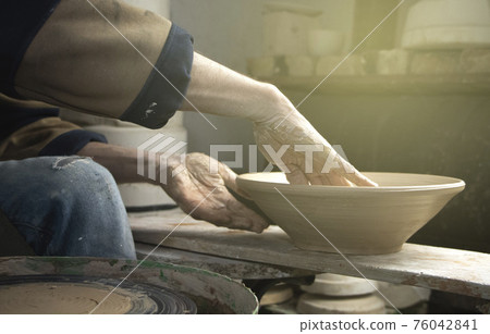 Hands of a potter on a pottery wheel close-up. Old traditional art, handmade, clay and ceramic production. Pottery workshop in subdued soft light Hands of a potter on a pottery wheel close-up. Old traditional art, handmade, clay and ceramic production. Pottery workshop in subdued soft light 76042841