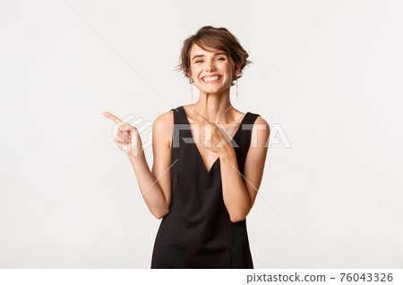 Cheerful stylish woman in black dress pointing fingers left, showing logo and smiling happy 76043326