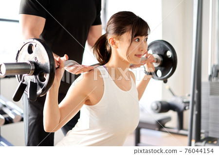 Asian woman doing barbell squats with the assistance of a trainer Asian woman doing barbell squats with the assistance of a trainer 76044154