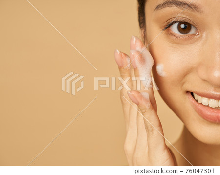 Studio half face portrait of beautiful young woman smiling at camera while applying gentle foam facial cleanser isolated over beige background 76047301