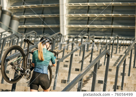 Rear view of female cyclist with bicycle on her shoulder walking up steps, having training in city Rear view of female cyclist with bicycle on her shoulder walking up steps, having training in city 76056499