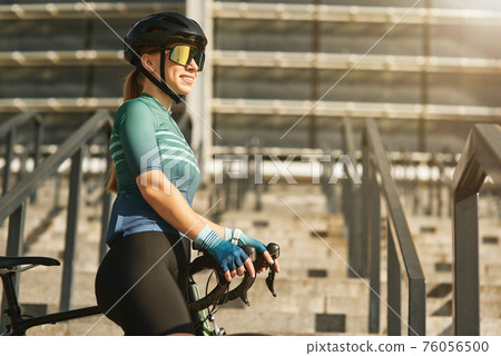 Sportive young woman, professional cyclist in cycling garment and protective gear smiling aside, standing on the steps with her bike while training outdoors Sportive young woman, professional cyclist in cycling garment and protective gear smiling aside, standing on the steps with her bike while training outdoors 76056500
