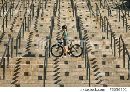 Young professional female cyclist in cycling garment and protective gear standing on the steps with her bike after having a training, riding in city Young professional female cyclist in cycling garment and protective gear standing on the steps with her bike after having a training, riding in city 76056501